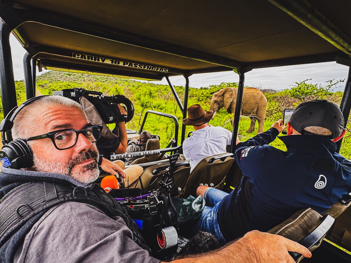 Wayne McKelvie in headphones with his sound kit in a safari vehicle, crew filming an elephant behind him